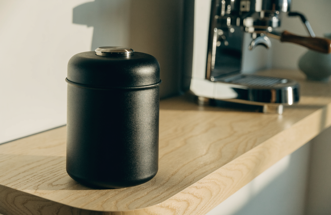 Matte black vacuum coffee canister on a pale oak counter beside an espresso machine