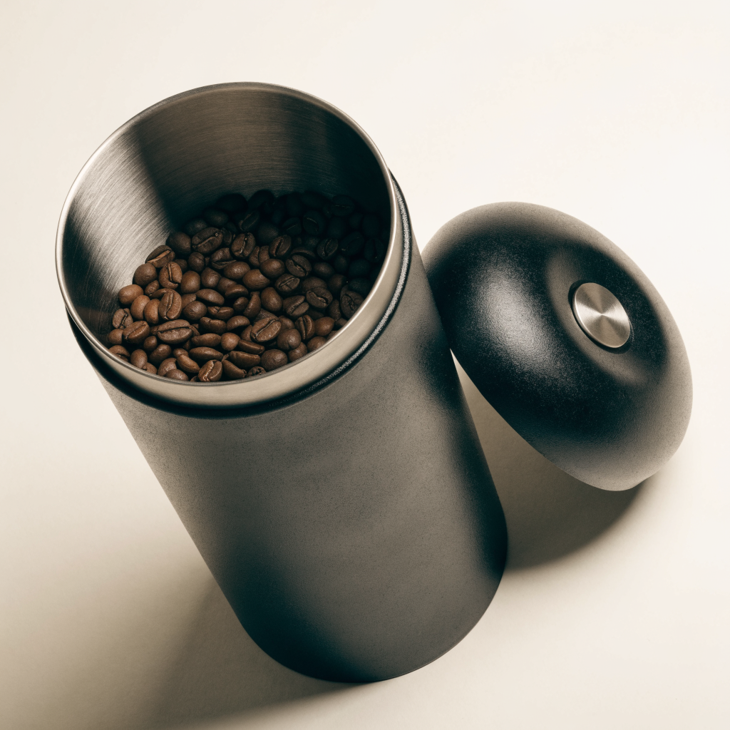 Vacuum coffee canister with the lid removed, showing the brushed stainless interior filled with coffee beans and the press-button dome lid resting beside it